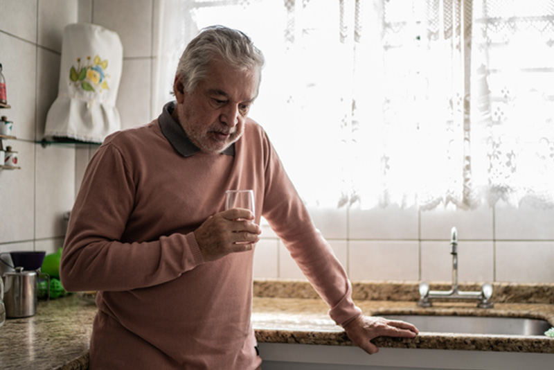 A sad senior man stands in the kitchen with a glass of water pondering the connection between heart disease and depression.